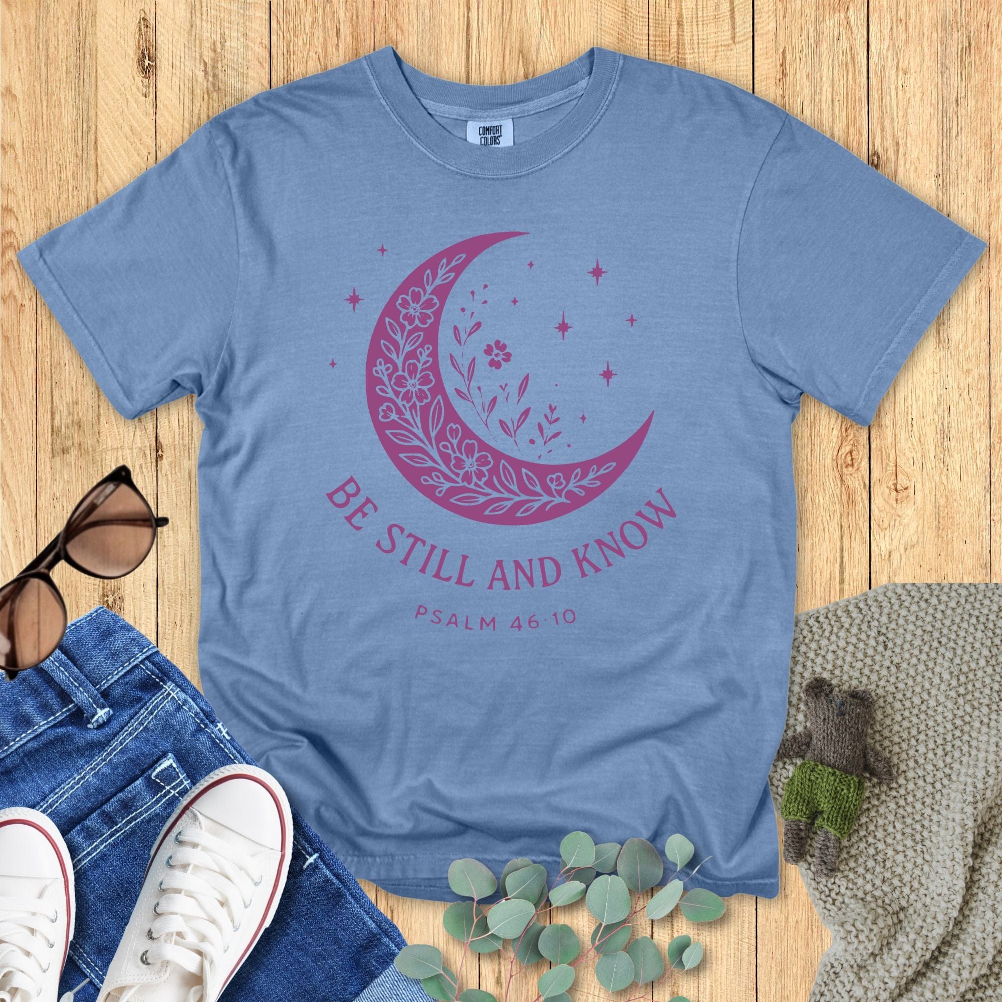 Flat lay of a Comfort Colors Christian T-shirt in blue jean, featuring a purple crescent moon filled with floral details and stars, with text “Be Still and Know – Psalm 46:10.”