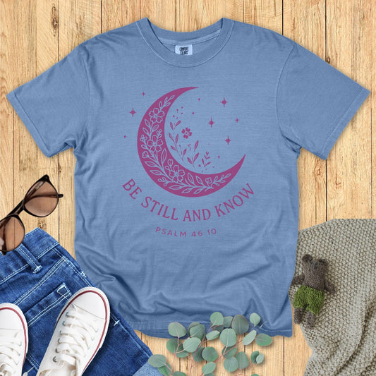 Flat lay of a Comfort Colors Christian T-shirt in blue jean, featuring a purple crescent moon filled with floral details and stars, with text “Be Still and Know – Psalm 46:10.”