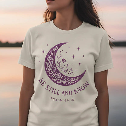 Woman wearing an ivory Comfort Colors Christian T-shirt with a purple crescent moon and floral starry design, displaying the scripture “Be Still and Know – Psalm 46:10,” photographed outdoors at sunset