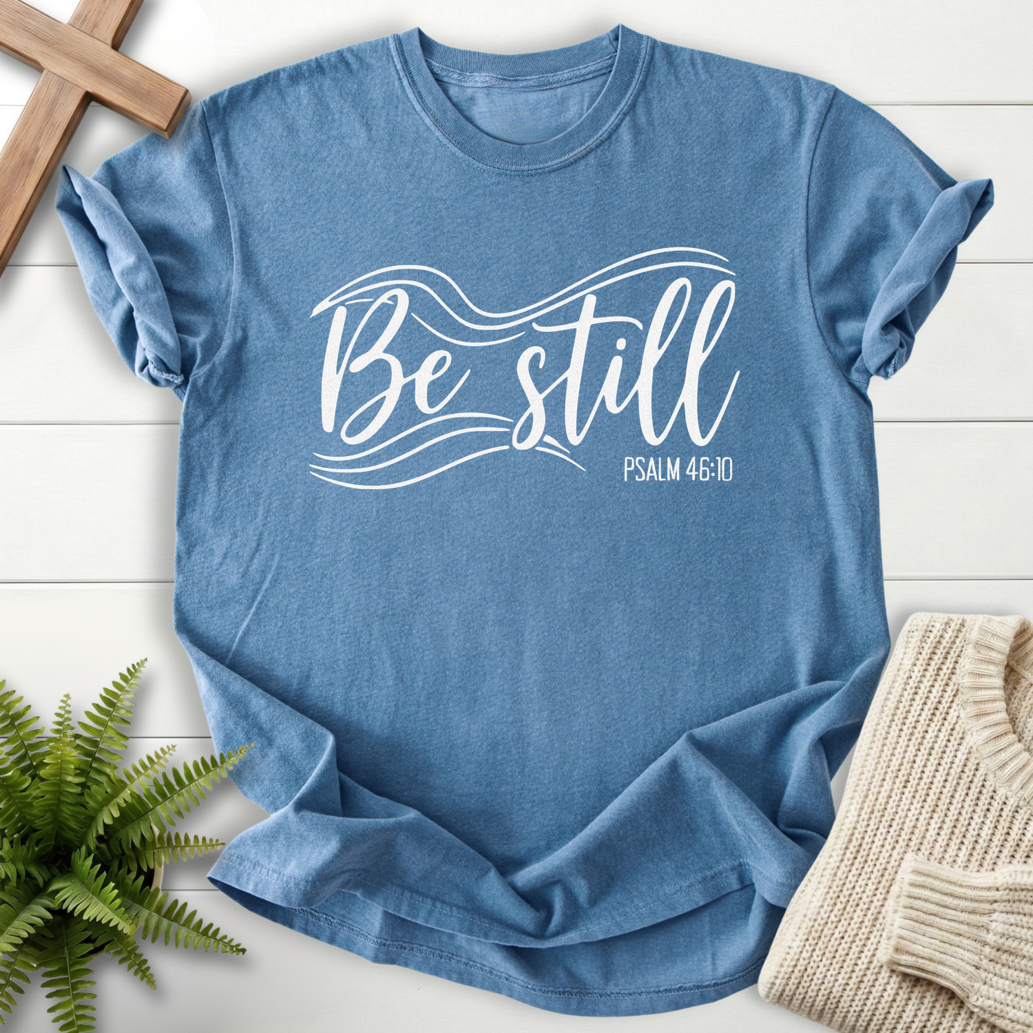Blue Jean Christian t-shirt for women with flowing white script lettering and wave accents, inspired by Psalm 46:10.