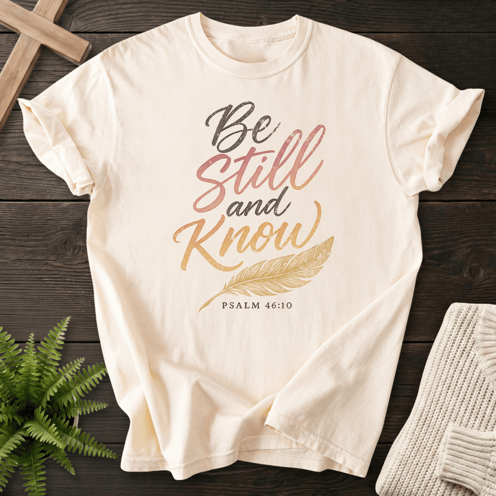 Ivory Christian t-shirt for women with gradient pink-to-gold 'Be Still and Know' script, gold feather illustration, and Psalm 46:10, She Wears Faith