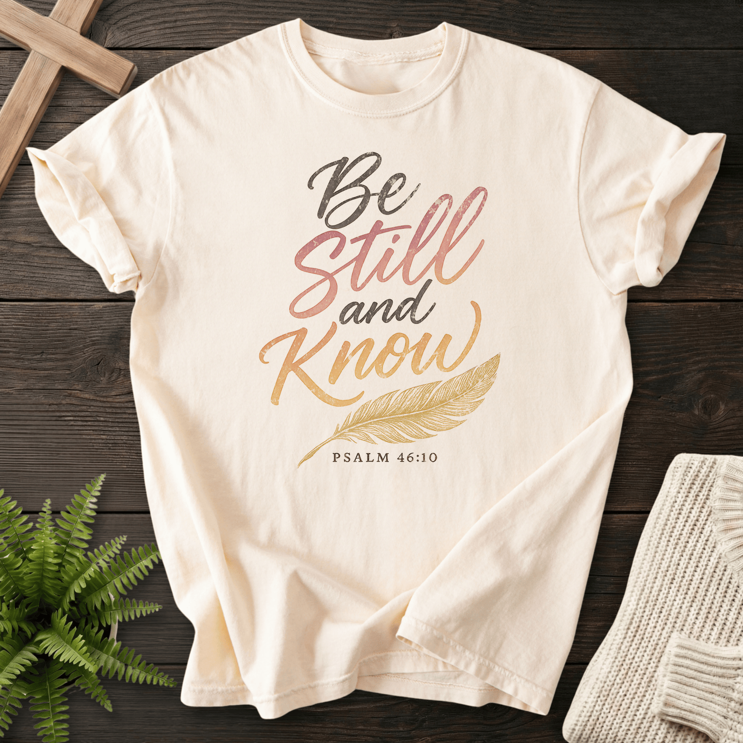 Ivory Christian t-shirt for women with gradient pink-to-gold 'Be Still and Know' script, gold feather illustration, and Psalm 46:10, She Wears Faith