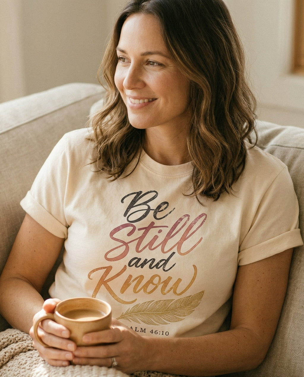 Be Still Feather Script – Ivory women's Christian tee, lifestyle photo in cozy living room – Gradient multicolor script lettering 'Be Still and Know' in pink – Psalm 46:10 – She Wears Faith