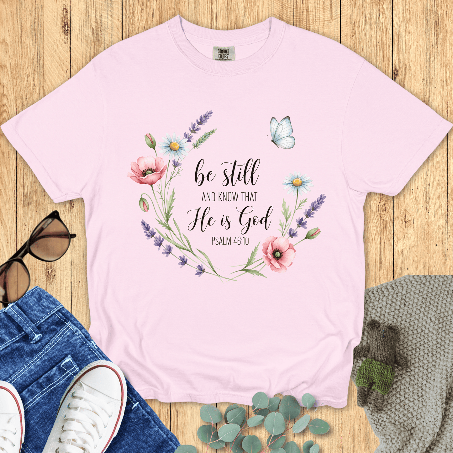 Blossom pink Comfort Colors Christian T-shirt with watercolor floral wreath and butterfly design, scripture "Be Still and Know That He Is God, Psalm 46:10." Religious inspirational tee flat lay with jeans, sneakers, and accessories.