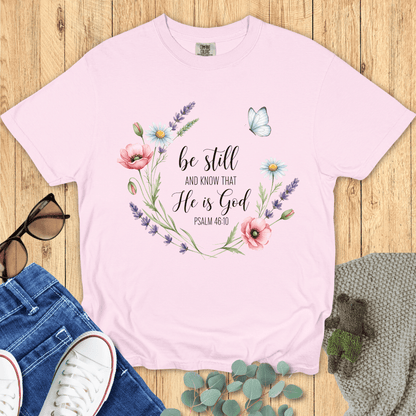 Blossom pink Comfort Colors Christian T-shirt with watercolor floral wreath and butterfly design, scripture "Be Still and Know That He Is God, Psalm 46:10." Religious inspirational tee flat lay with jeans, sneakers, and accessories.