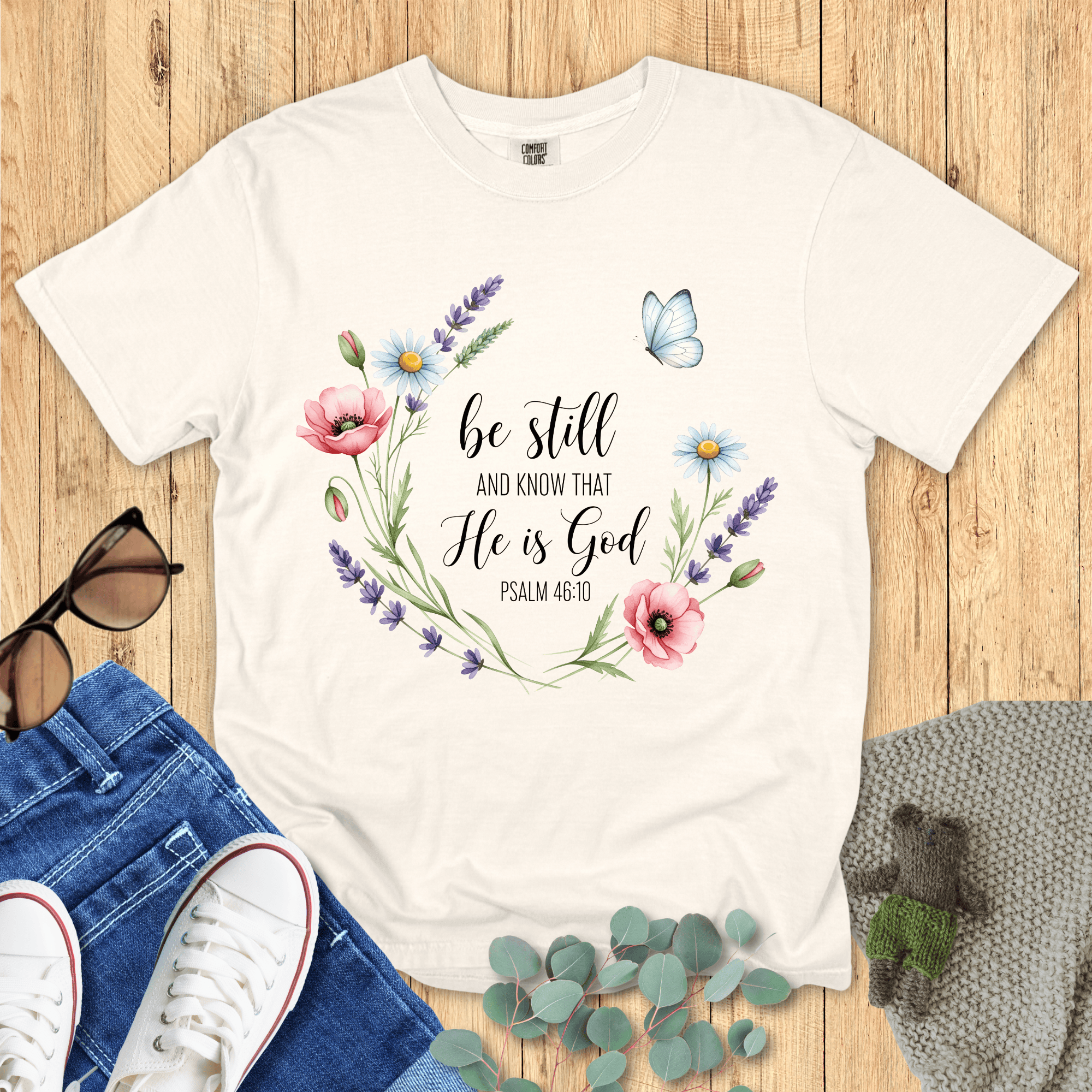Ivory Comfort Colors Christian T-shirt with floral wreath and butterfly design featuring scripture "Be Still and Know That He Is God, Psalm 46:10." Inspirational Christian tee flat lay with jeans, sneakers, and sunglasses on wood background.
