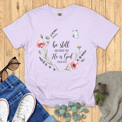 Orchid purple Comfort Colors Christian T-shirt with floral wreath and butterfly artwork around verse "Be Still and Know That He Is God, Psalm 46:10." Faith-based graphic tee flat lay with casual outfit on wooden surface.