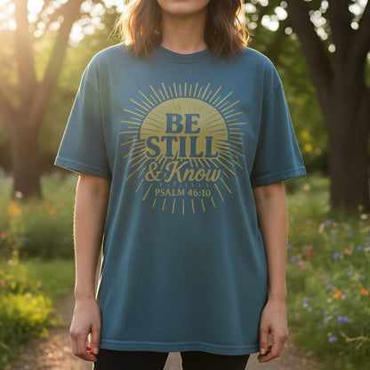 Blue Spruce Comfort Colors Christian T-shirt with golden sunburst graphic and scripture "Be Still & Know, Psalm 46:10." Modeled outdoors in a sunlit garden for a natural lifestyle look.