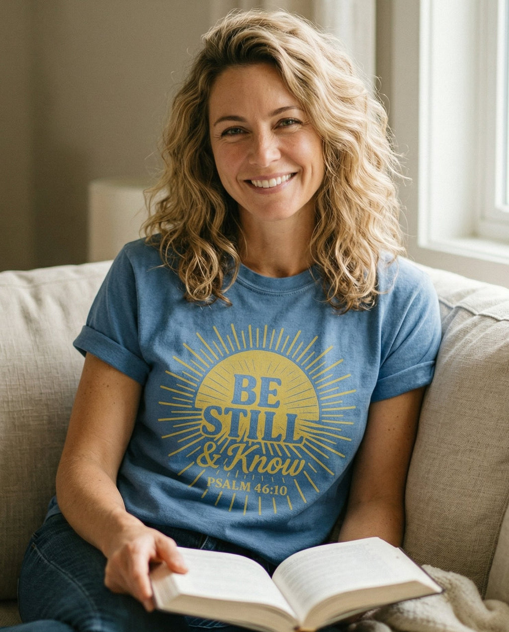 Be Still & Know Sunburst – Blue Jean women's Christian tee, lifestyle photo in cozy living room – Bold sunburst radiating graphic with 'BE STILL & Know' lettering insid – Psalm 46:10 – She Wears Faith