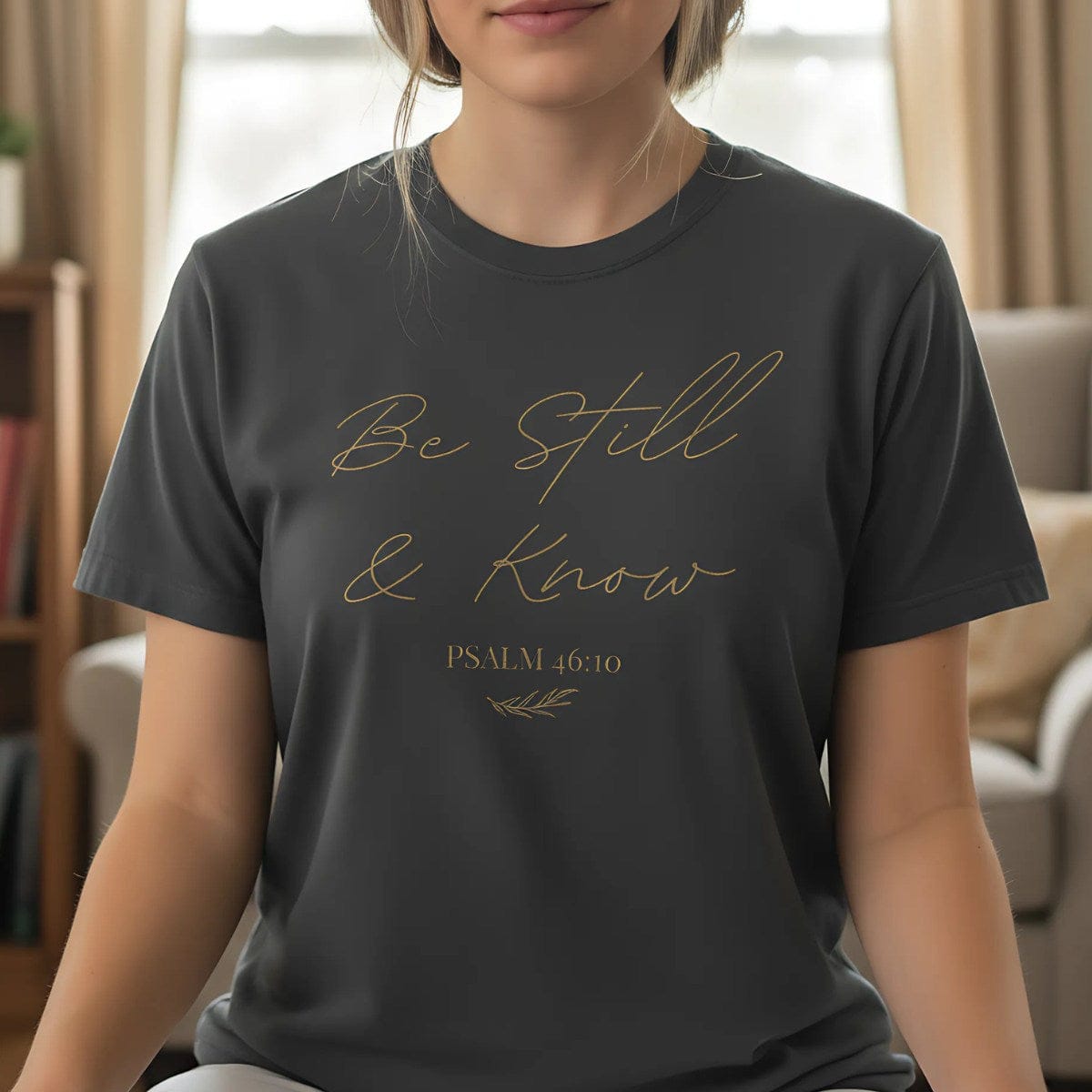 Woman wearing a dark gray T-shirt with the phrase "Be Still & Know" and Psalm 46:10 in script, sitting peacefully indoors, suggesting calm and meditation