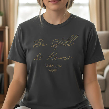 Woman wearing a dark gray T-shirt with the phrase "Be Still & Know" and Psalm 46:10 in script, sitting peacefully indoors, suggesting calm and meditation
