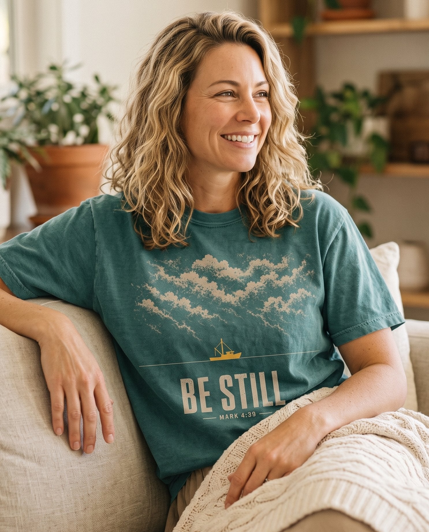 Be Still Mark 4:39 – Blue Spruce women's Christian tee, lifestyle photo in cozy living room – Tan storm clouds rolling across the top with a small golden fishing bo – Mark 4:39 – She Wears Faith