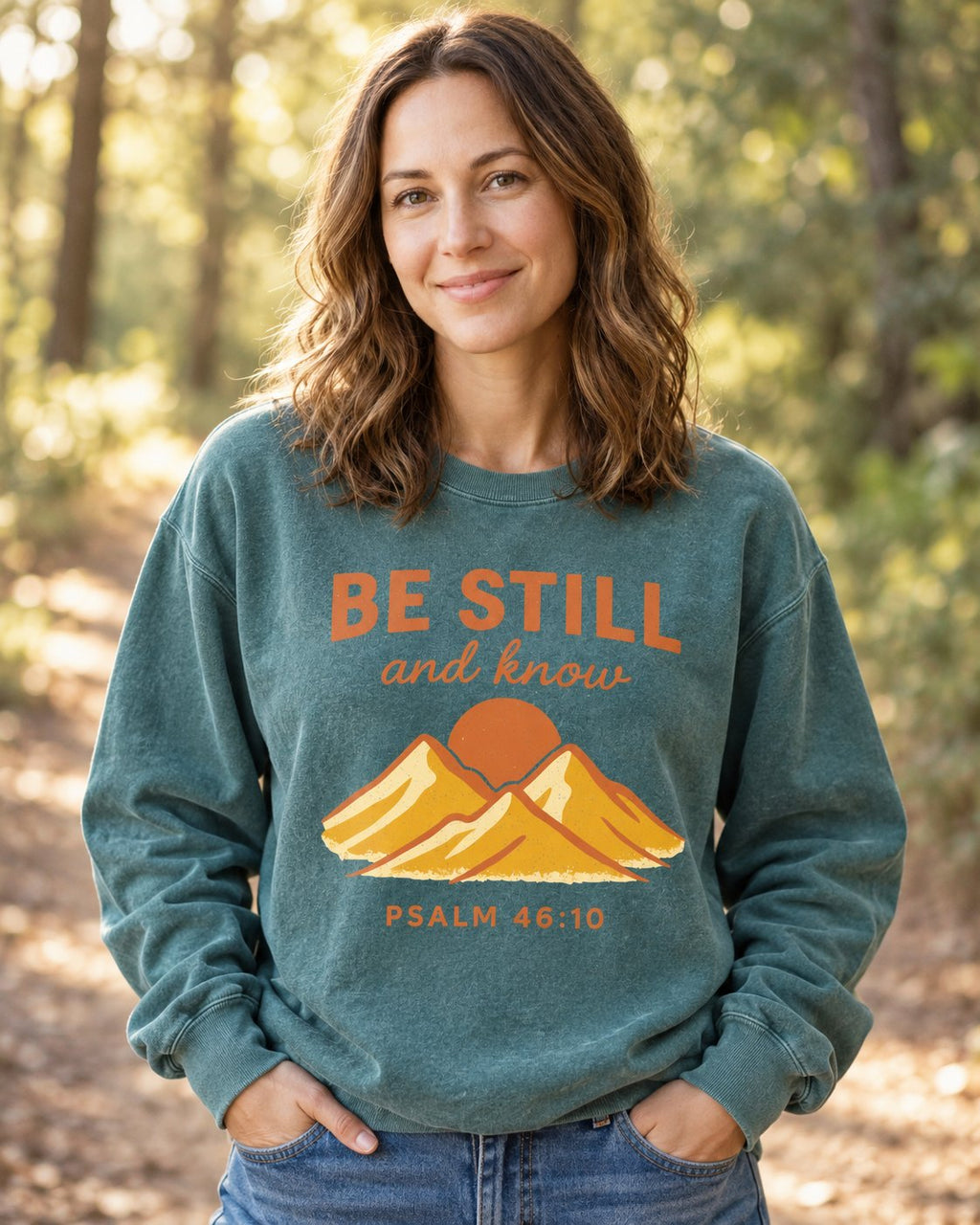 Be Still Mountain Sunrise – Blue Spruce women's Christian sweatshirt, lifestyle photo in nature trail – Orange BE STILL over gold mountains and a sun – She Wears Faith