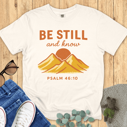 Ivory Comfort Colors Christian T-shirt with retro mountain and sunrise graphic, featuring Bible verse "Be Still and Know, Psalm 46:10." Inspirational faith-based tee flat lay with jeans, sneakers, and sunglasses on wood background.