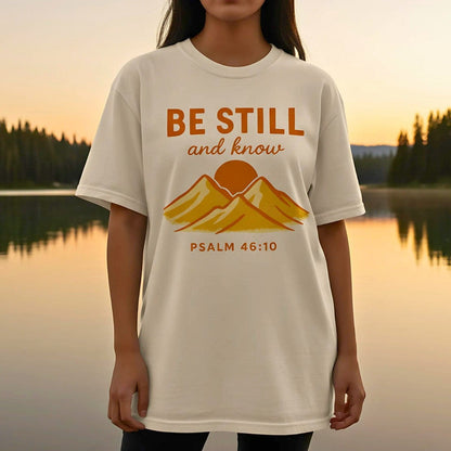 Ivory Comfort Colors Christian T-shirt with retro mountain and sun design and scripture "Be Still and Know, Psalm 46:10." Modeled outdoors at sunset near a lake for a natural lifestyle look.