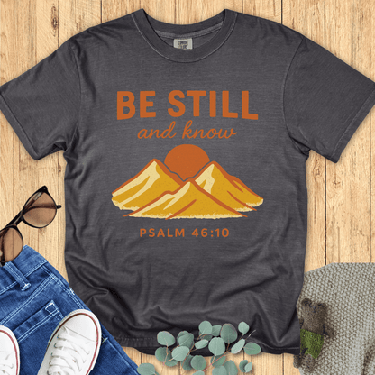 Pepper gray Comfort Colors Christian T-shirt with golden mountain illustration and verse "Be Still and Know, Psalm 46:10." Religious inspirational tee flat lay with casual outfit pieces on wooden background.