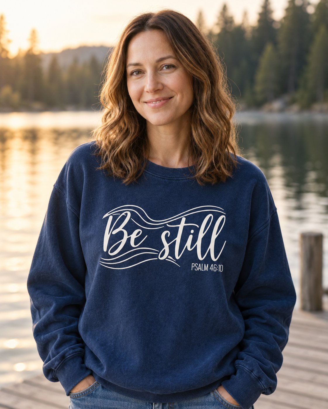 Be Still – True Navy women's Christian sweatshirt, lifestyle photo in lakeside setting – White Be still script with flowing wave lines – She Wears Faith