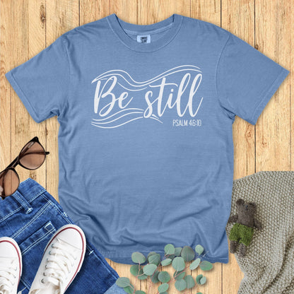 Blue jean Christian T-shirt flat lay featuring the phrase “Be Still” with flowing white wave accents and “Psalm 46:10.” Displayed with sunglasses, denim jeans, sneakers, eucalyptus leaves, and a knitted blanket on a wooden background