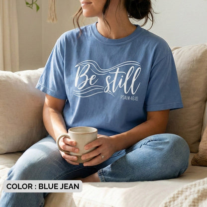 Blue jean Christian Women's T-Shirt showing the words "Be still" in large white script with decorative wave lines and the verse reference "Psalm 46:10".