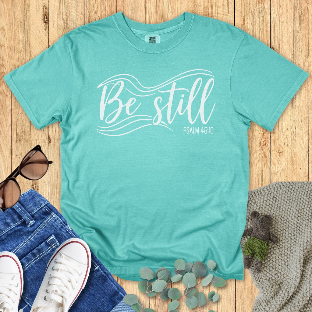 Seafoam green Christian T-shirt flat lay with the phrase “Be Still” in elegant white lettering and flowing wave graphics, alongside “Psalm 46:10.” Styled with sunglasses, jeans, sneakers, eucalyptus leaves, and a knitted blanket on a wooden background