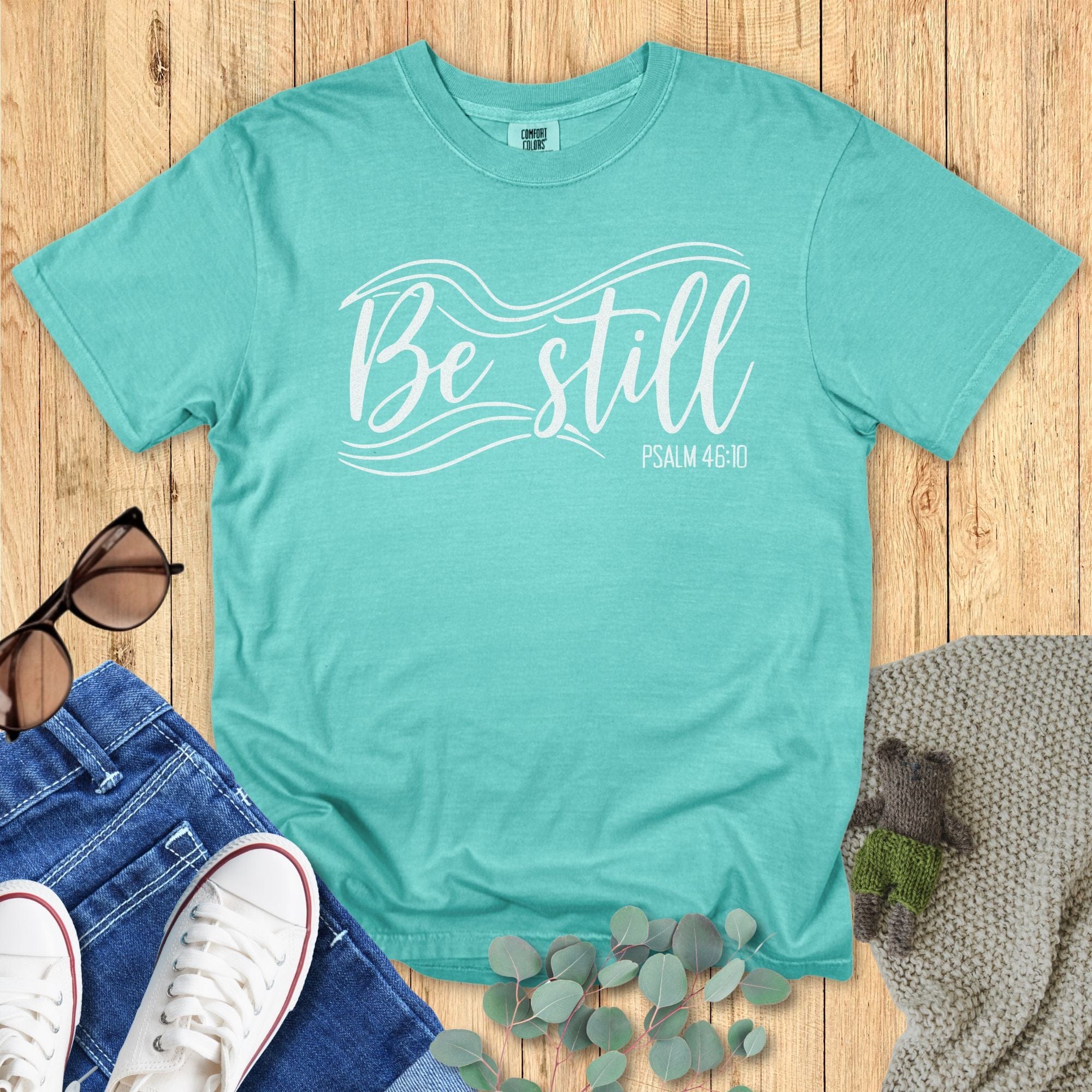 Seafoam green Christian T-shirt flat lay with the phrase “Be Still” in elegant white lettering and flowing wave graphics, alongside “Psalm 46:10.” Styled with sunglasses, jeans, sneakers, eucalyptus leaves, and a knitted blanket on a wooden background