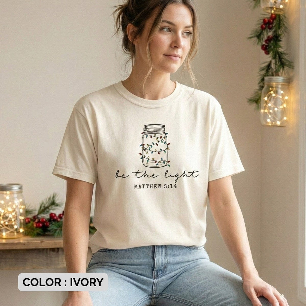 Christian women t-shirt worn by a model in ivory Comfort Colors 1717, featuring a mason jar light illustration and “Be the Light” Matthew 5:14 verse, soft casual US faith wear style.