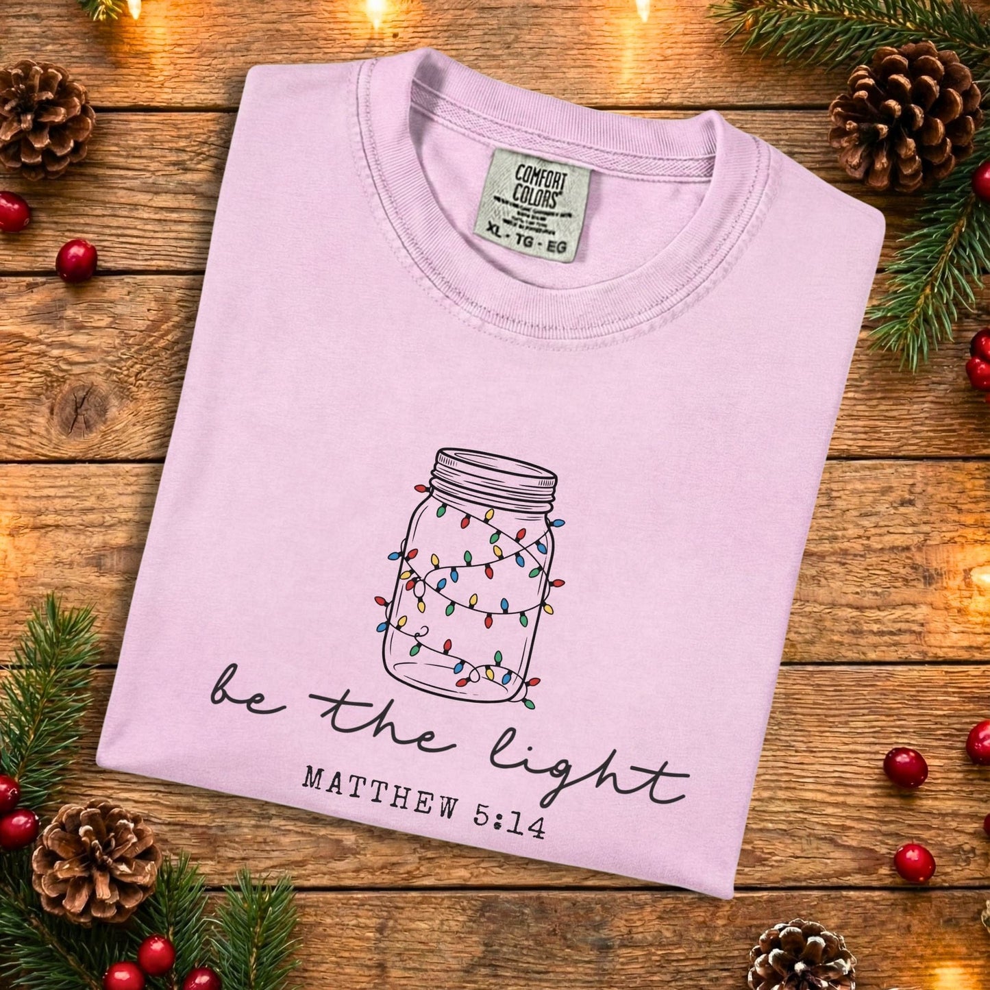 Blossom pink Comfort Colors 1717 Christian women tee with a hand-drawn mason jar wrapped in colorful Christmas lights and the phrase “be the light” Matthew 5:14, folded flat on a rustic wood background, soft US faith wear t-shirt.