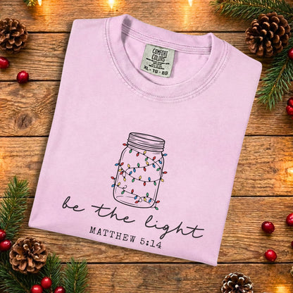 Blossom pink Comfort Colors 1717 Christian women tee with a hand-drawn mason jar wrapped in colorful Christmas lights and the phrase “be the light” Matthew 5:14, folded flat on a rustic wood background, soft US faith wear t-shirt.