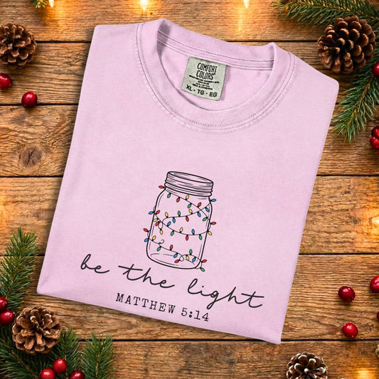 Blossom pink Comfort Colors 1717 Christian women tee with a hand-drawn mason jar wrapped in colorful Christmas lights and the phrase “be the light” Matthew 5:14, folded flat on a rustic wood background, soft US faith wear t-shirt.