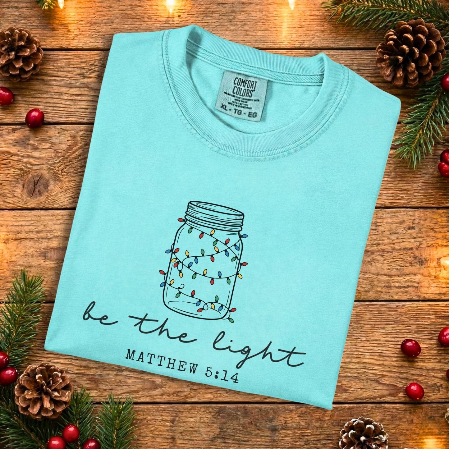Chalky mint Comfort Colors 1717 Christian women tee with a mason jar light illustration and “Be the Light” Matthew 5:14 scripture, laid flat on a rustic background, faith-inspired US faith wear t-shirt.