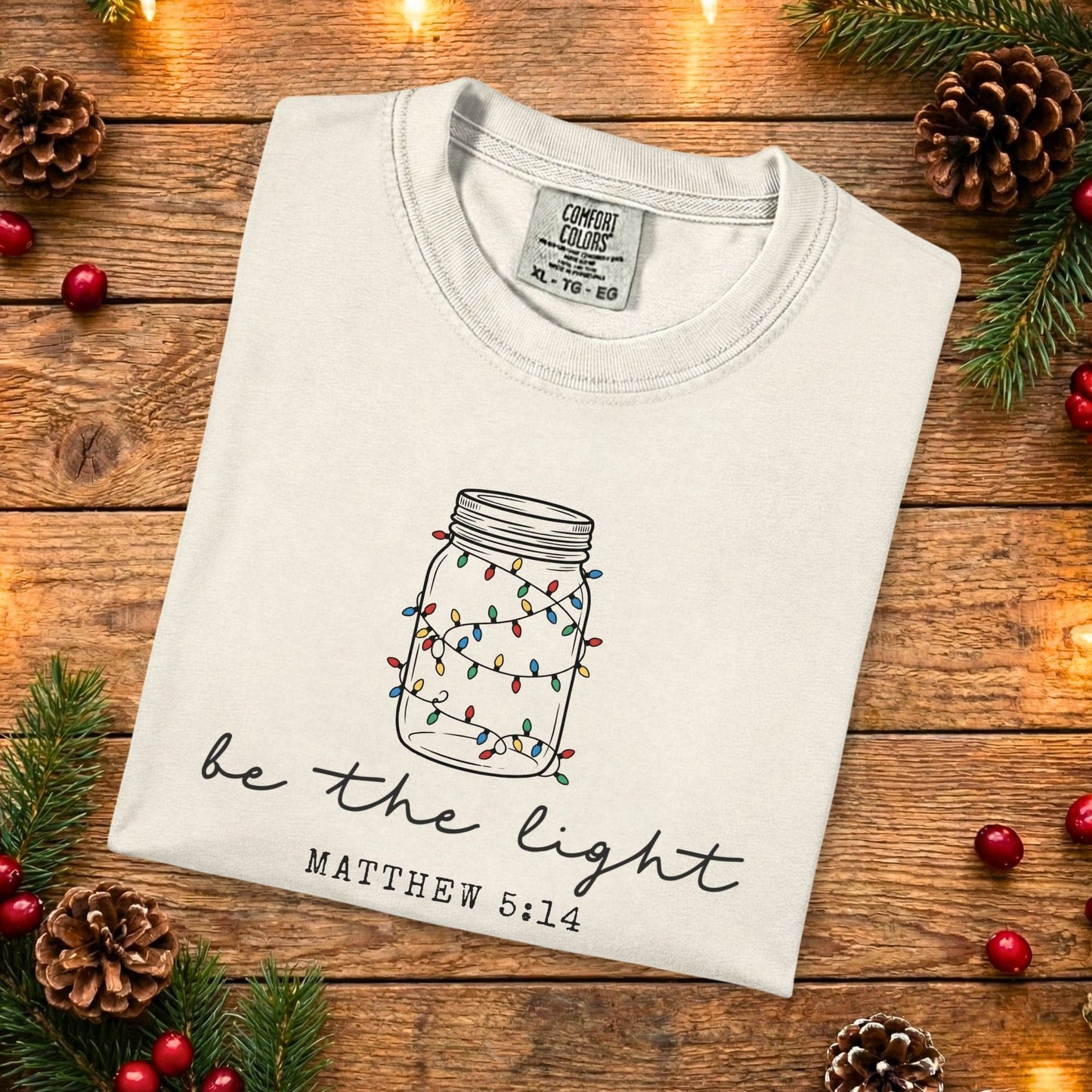 Ivory Comfort Colors 1717 Christian women tee featuring a hand-drawn mason jar with colorful lights and the scripture “Be the Light” Matthew 5:14, folded flat on a rustic wood background, faith-based US faith wear t-shirt.