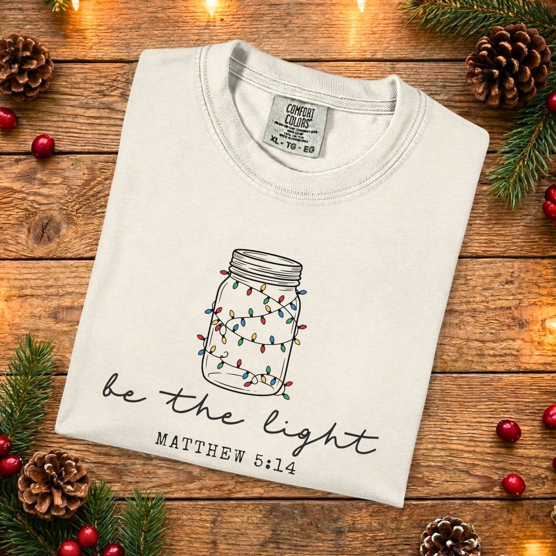Ivory Comfort Colors 1717 Christian women tee featuring a hand-drawn mason jar with colorful lights and the scripture “Be the Light” Matthew 5:14, folded flat on a rustic wood background, faith-based US faith wear t-shirt.