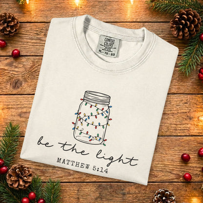 Ivory Comfort Colors 1717 Christian women tee featuring a hand-drawn mason jar with colorful lights and the scripture “Be the Light” Matthew 5:14, folded flat on a rustic wood background, faith-based US faith wear t-shirt.