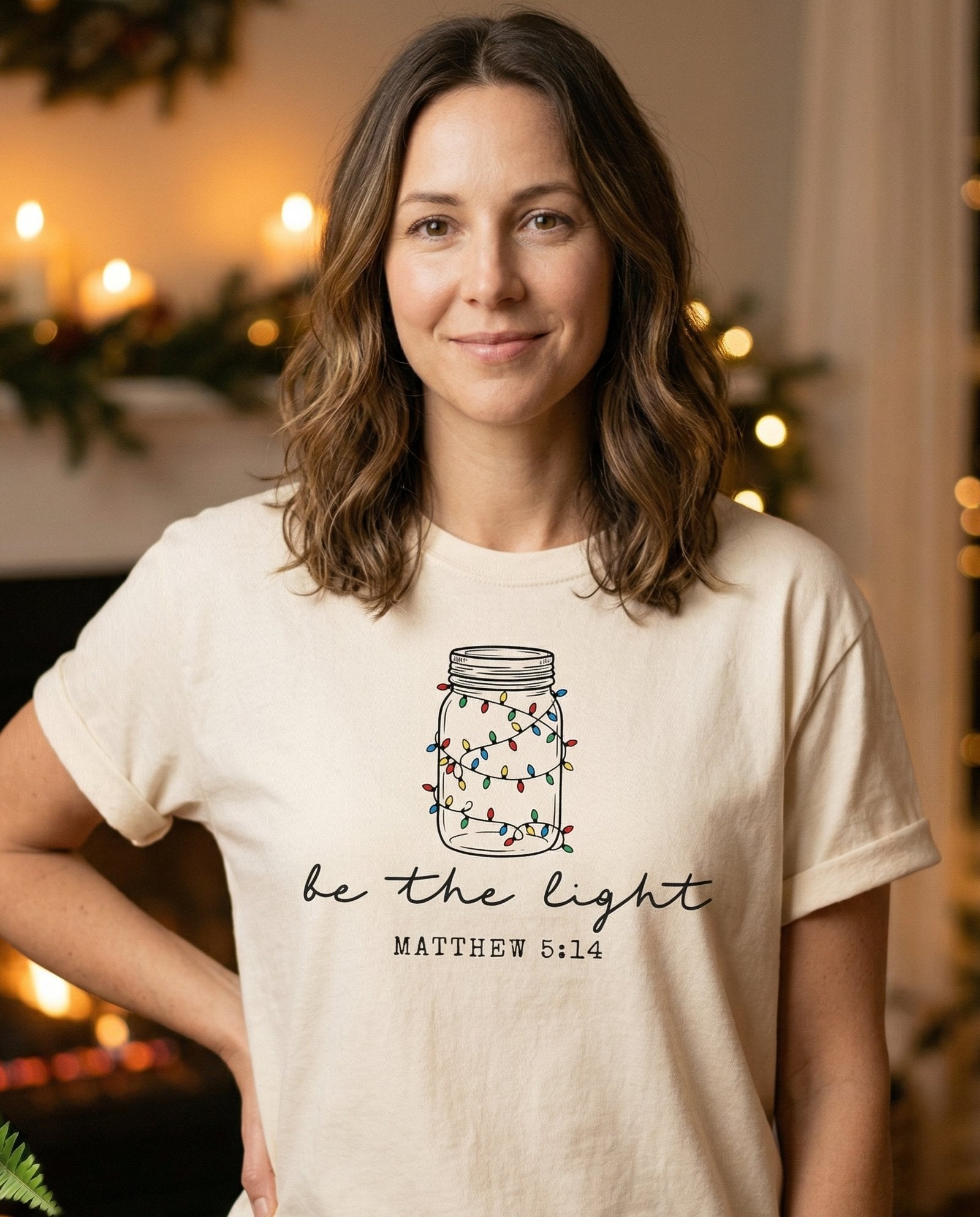 Be the Light Mason Jar – Ivory women's Christian tee, lifestyle photo in cozy holiday setting – Line art illustration of a mason jar filled with colorful Christmas li – Matthew 5:14 – She Wears Faith