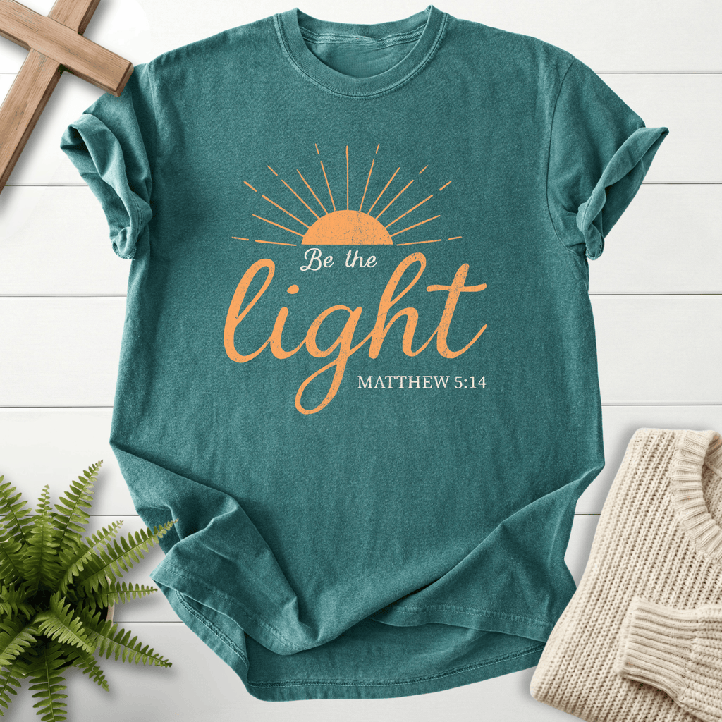 Blue Spruce Christian t-shirt for women with a sunrise illustration and script lettering, 'Be the Light', inspired by Matthew 5:14.