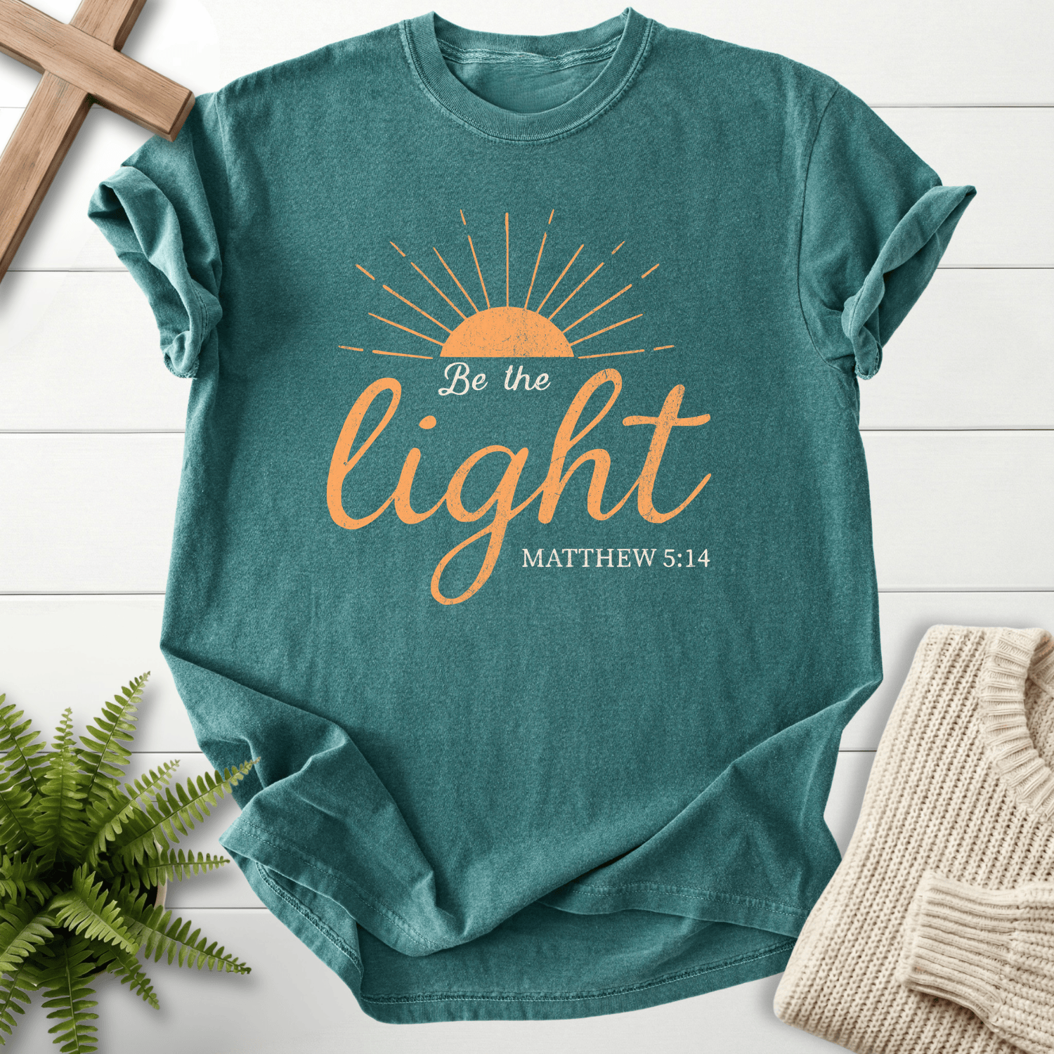 Blue Spruce Christian t-shirt for women with a sunrise illustration and script lettering, 'Be the Light', inspired by Matthew 5:14.