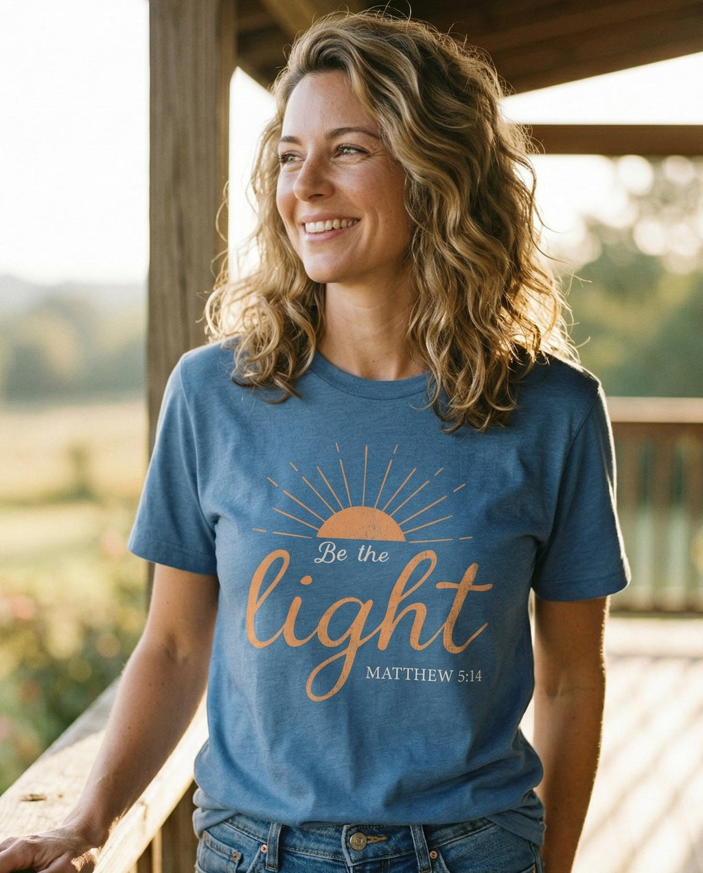 Be the Light – Blue Jean women's Christian tee, lifestyle photo in sunny porch – Half-sun rising with rays illustration above elegant script 'Be the li – Matthew 5:14 – She Wears Faith