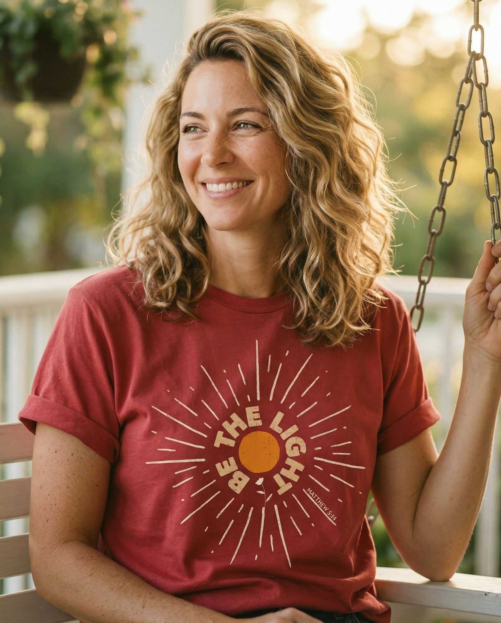 Be The Light Sunburst T-shirt – Crimson women's Christian tee, lifestyle photo in sunny porch – Bold circular sunburst with 'BE THE LIGHT' text following the arc – Matthew 5:14 – She Wears Faith