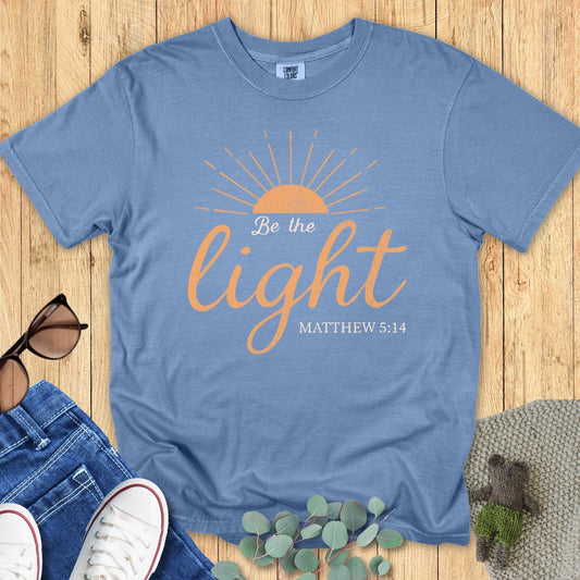 Comfort Colors 1717 blue jean “Be the Light – Matthew 5:14” t-shirt flatlay on a wooden background with denim and sneakers. A modern Christian shirt from women’s scripture tees collection.