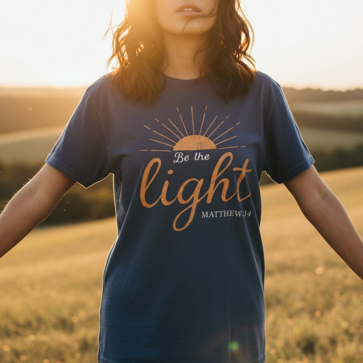 Woman standing in a golden field at sunset wearing a Comfort Colors 1717 navy “Be the Light – Matthew 5:14” t-shirt. A faith-based apparel for women design inspired by Matthew 5:14 t-shirt message.