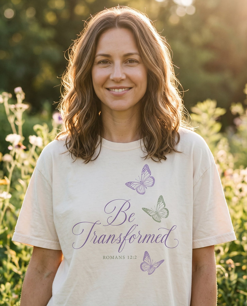 Be Transformed Romans 12:2 – Ivory women's Christian tee, lifestyle photo in outdoor garden – Purple cursive lettering 'Be Transformed' with ROMANS 12:2 in sage gre – Romans 12:2 – She Wears Faith