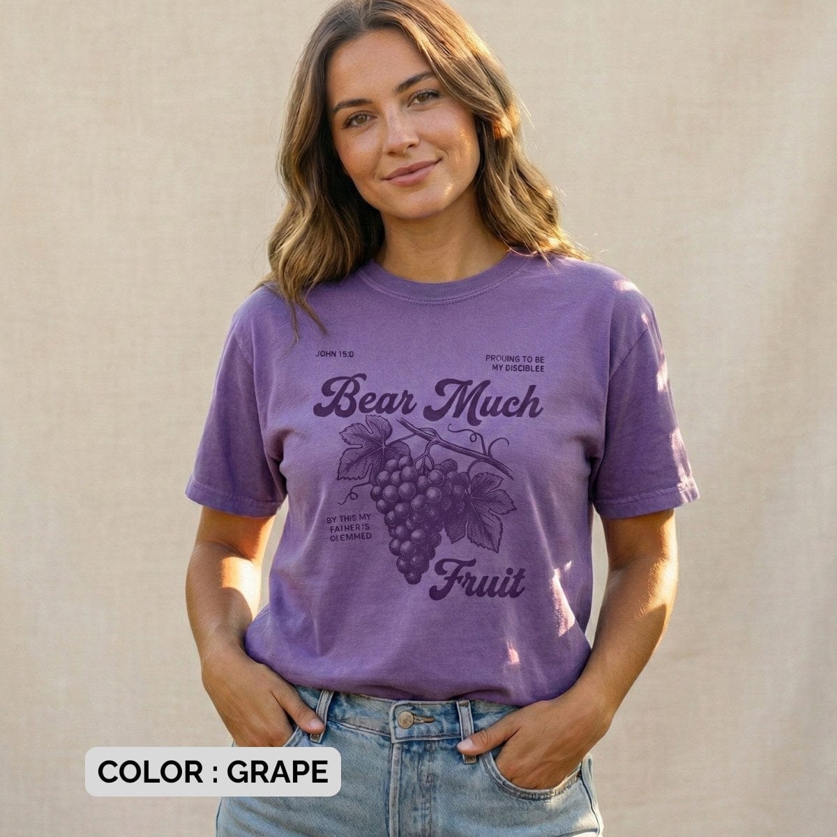 Christian women tee in Grape, Comfort Colors 1717, featuring the “Bear Much Fruit” John 15:8 grape illustration; model wearing US faith wear outdoors against a soft neutral backdrop, hands in pockets, relaxed and joyful style.