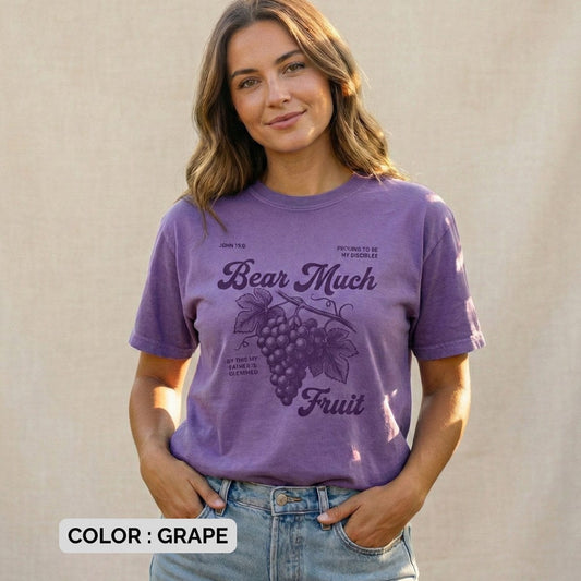 Christian women tee in Grape, Comfort Colors 1717, featuring the “Bear Much Fruit” John 15:8 grape illustration; model wearing US faith wear outdoors against a soft neutral backdrop, hands in pockets, relaxed and joyful style.