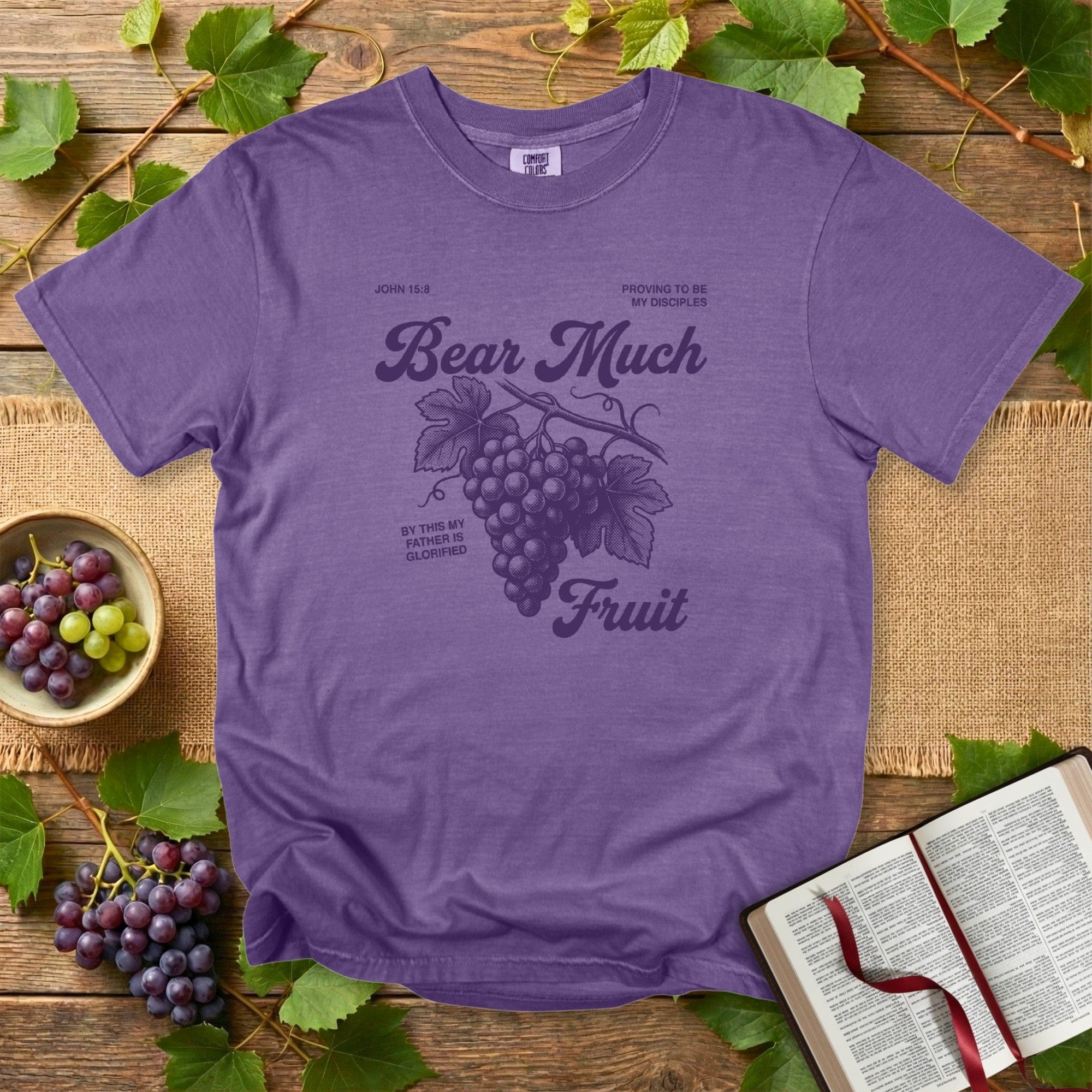Grape-colored Christian women tee on Comfort Colors 1717 showcasing the “Bear Much Fruit” John 15:8 artwork with hand-drawn grapes, set against a rustic wood background for US faith wear branding.