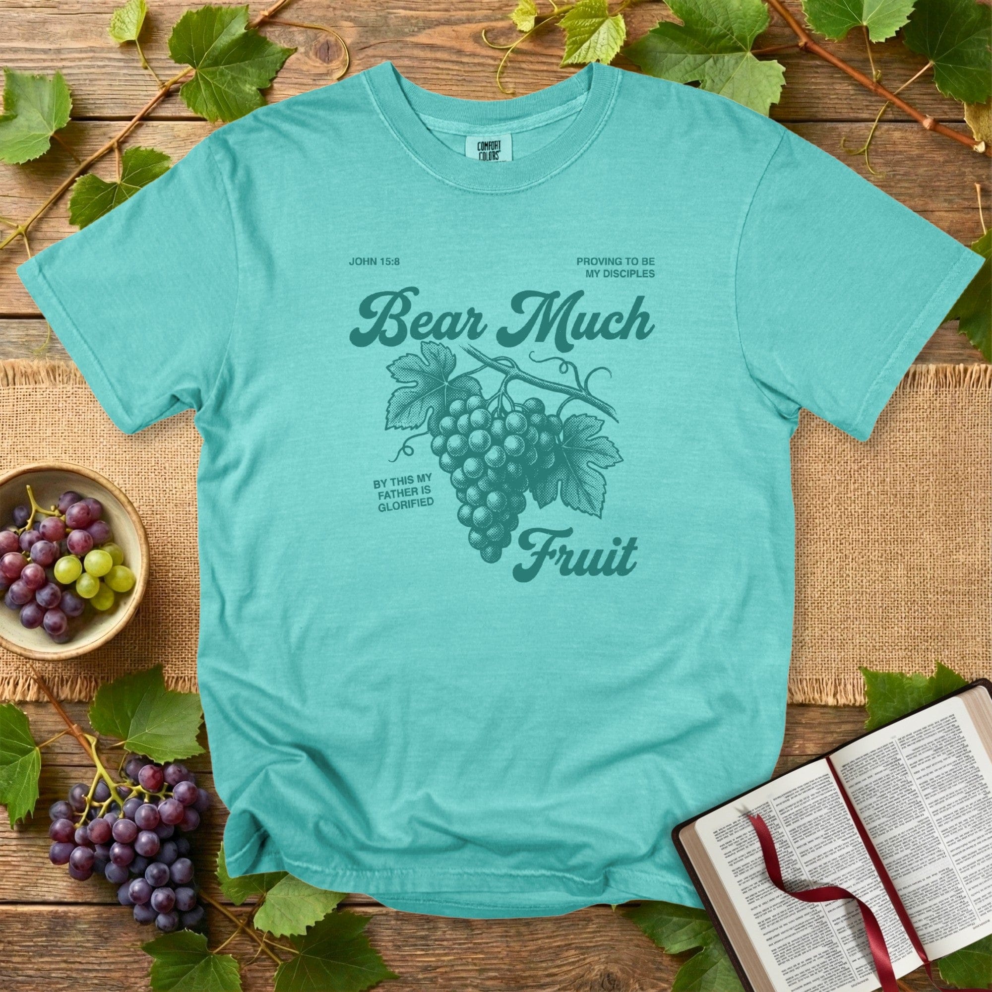 Seafoam Comfort Colors 1717 Christian women tee featuring “Bear Much Fruit” from John 15:8 with a vintage grape illustration, styled on a rustic table with grapes and an open Bible; modern US faith wear.