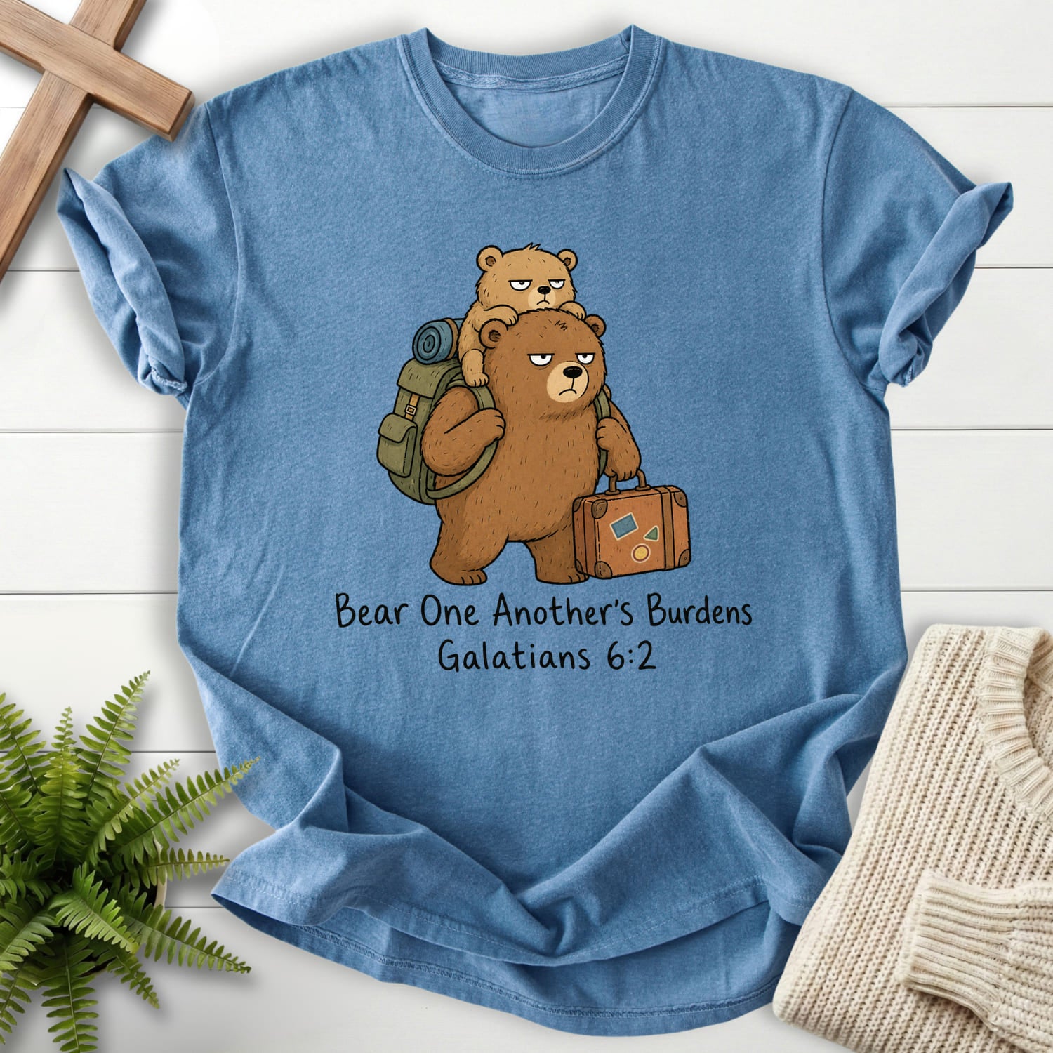Flatlay of a Blue Jean women's Christian tee with "Bear One Another's Burdens T-Shirt" Galatians 6:2.