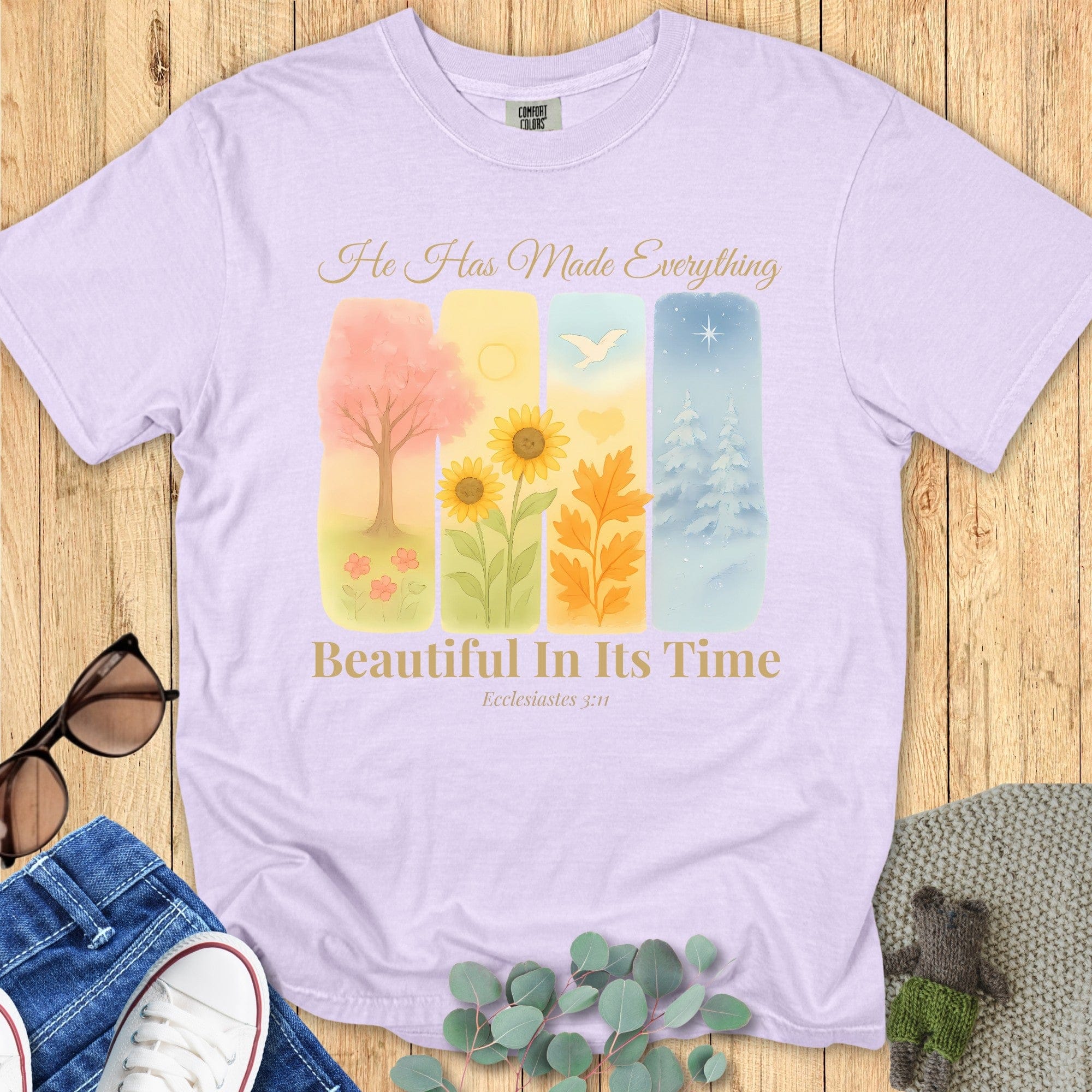 Flatlay of a Comfort Colors 1717 orchid “He Has Made Everything Beautiful in Its Time – Ecclesiastes 3:11” t-shirt featuring a soft watercolor design of four seasons. A feminine and uplifting women’s scripture tee and faith-based apparel for women.