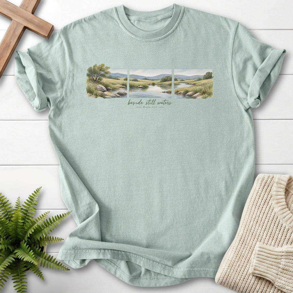 Flat lay of a Bay Scripture shirt featuring watercolor artwork with "Beside Still Waters T-Shirt" Psalm 23:2.