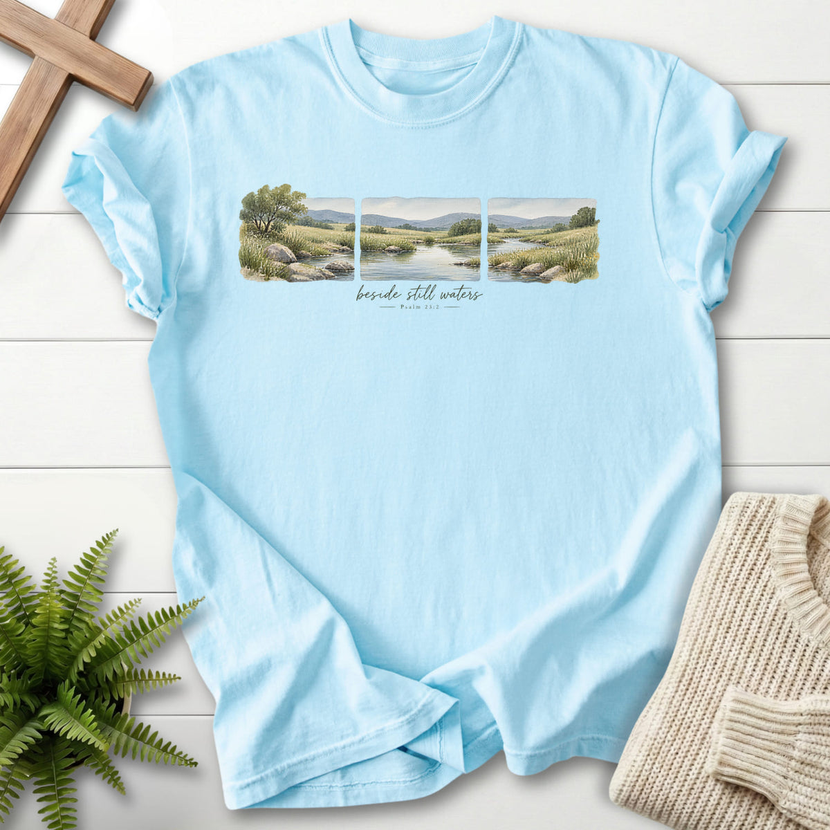 Flatlay of a Chambray Bible verse tee with watercolor artwork with "Beside Still Waters T-Shirt" Psalm 23:2.