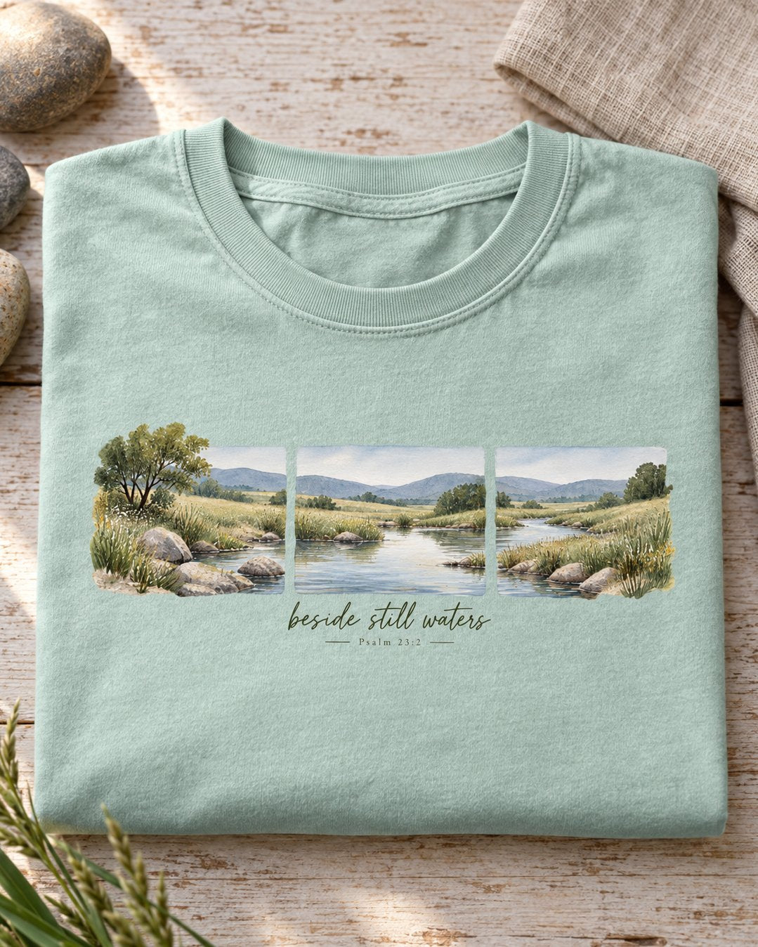 Beside Still Waters – Bay women's Christian tee, folded flat lay – Three watercolor panels of a quiet stream through green hills – Psalm 23:2 – She Wears Faith